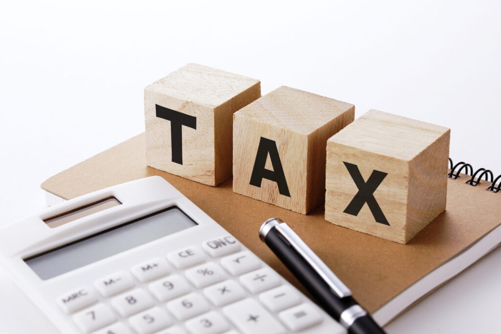 電卓とボールペンが並び置かれている写真。ノートの上には"TAX"と書かれた立方体の積み木が載っている。税金に関した記事内容を表現。